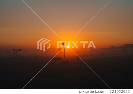 A tall, abstract art installation resembling a human figure stands silhouetted against a vibrant desert sunset, with festival participants in the distance. A tall, abstract art installation resembling a human figure stands silhouetted against a vibrant desert sunset, with festival participants in the distance. 121952633