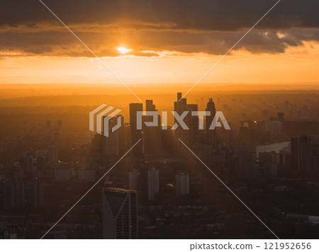 The London skyline is silhouetted against a vibrant sunset, with the River Thames winding through the cityscape, creating a serene atmosphere. 121952656