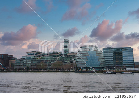 The iconic London City Hall's glass structure is set against a partly cloudy sky, flanked by modern buildings, with the River Thames reflecting the scene. 121952657
