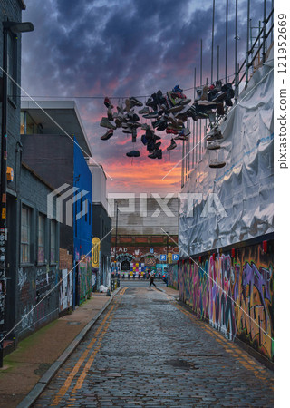 A narrow London street features vibrant graffiti and a unique display of hanging shoes, set against a dramatic sunset sky, capturing an urban artistic vibe. 121952669
