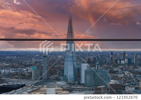 The Shard stands prominently in London's skyline at sunset, with a faint rainbow in the sky. Surrounding buildings highlight the city's bustling nature. 121952670