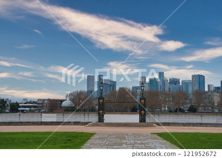 View of London's skyline featuring Canary Wharf skyscrapers, a decorative wrought iron gate, ornate lampposts, and a stone pathway by the Thames. 121952672