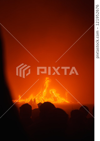 Silhouettes of people watch a large fire against a dark night sky, with intense orange and red flames at the Burning Man festival in Black Rock Desert. 121952676