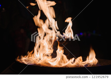 Flames dance around a skull shaped object in a fire pit, set against a dark background, highlighting the artistic installation at a desert festival. 121952679