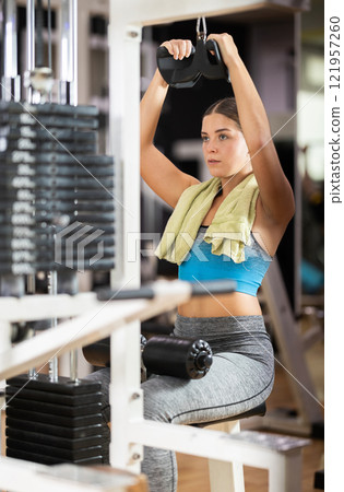 Girl in gym performs exercises for arm muscles using cable crossover machine and pull weighted rope 121957260