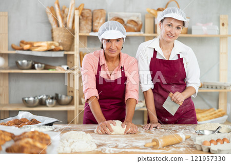 Experienced aged woman in apron working with young female assistant in bakery, kneading dough and preparing portioned pieces for baking 121957330
