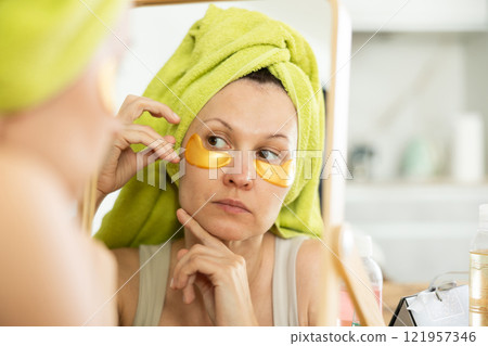 Middle-aged woman applying eye patch mask on her face sitting in front of the mirror 121957346