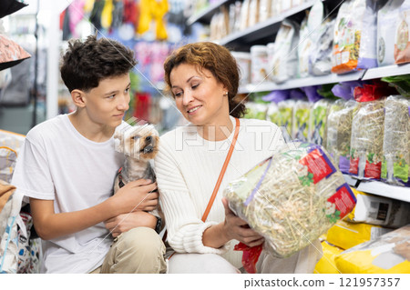Woman with teenage son and Yorkshire terrier choosing hay for rabbit cage in store 121957357