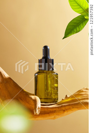 On a beige backdrop, a green amber glass bottle with a mockup of a serum is set in front of a piece of bleached driftwood, with some green leaves. An advertisement for a cosmetic product. On a beige backdrop, a green amber glass bottle with a mockup of a serum is set in front of a piece of bleached driftwood, with some green leaves. An advertisement for a cosmetic product. 121957392