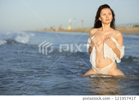 Portrait of woman in white panties and transparent turtleneck posing in sea waves in surf zone 121957417