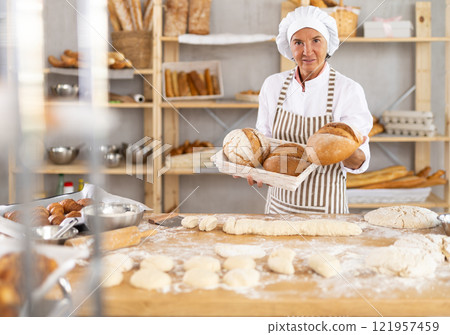 Successful senior female baker holding tray with fresh bread 121957459