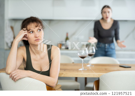 Upset middle-aged woman sitting at the kitchen table with her back to another lady quarreling to her Upset middle-aged woman sitting at the kitchen table with her back to another lady quarreling to her 121957521