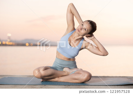 Near sea during sunset, girl performs variations of padmasana with body tilt 121957528