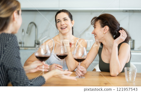 Three happy middle-aged women sitting at table with a glass of wine while having lively conversation Three happy middle-aged women sitting at table with a glass of wine while having lively conversation 121957543