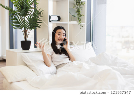 Asian woman in casual white shirt having phone call while relaxing on comfortable bed in bright room. Asian woman in casual white shirt having phone call while relaxing on comfortable bed in bright room. 121957547