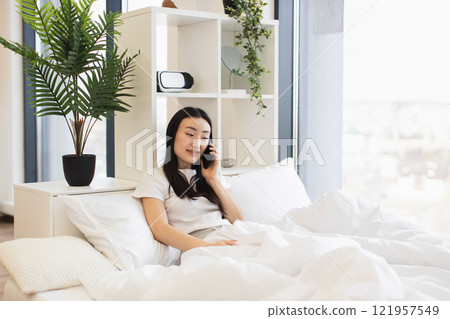 Asian woman in white room sitting on bed talking on phone, creating a calm and relaxed atmosphere. Asian woman in white room sitting on bed talking on phone, creating a calm and relaxed atmosphere. 121957549