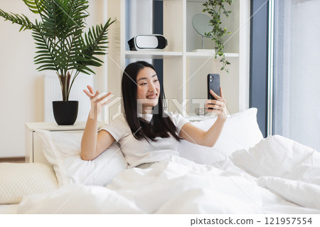 Asian woman in her 20s using smartphone for online video call while sitting on bed. Relaxed atmosphere with indoor plants and modern decor. Asian woman in her 20s using smartphone for online video call while sitting on bed. Relaxed atmosphere with indoor plants and modern decor. 121957554