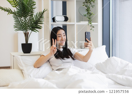 Asian woman in her 20s having video call on bed. She holds smartphone while smiling and making peace sign, conveying joy and connectivity during online chat. Asian woman in her 20s having video call on bed. She holds smartphone while smiling and making peace sign, conveying joy and connectivity during online chat. 121957556