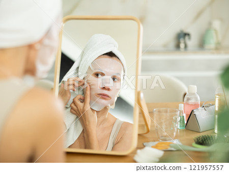 Woman applying moisturizing sheet face mask in front of mirror Woman applying moisturizing sheet face mask in front of mirror 121957557