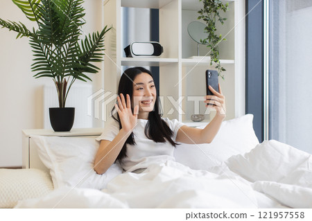 Young Asian woman having online video chat in cozy bedroom setting. She is smiling, holding smartphone, engaging in virtual communication, wearing casual attire. Bright, relaxing indoor environment. Young Asian woman having online video chat in cozy bedroom setting. She is smiling, holding smartphone, engaging in virtual communication, wearing casual attire. Bright, relaxing indoor environment. 121957558