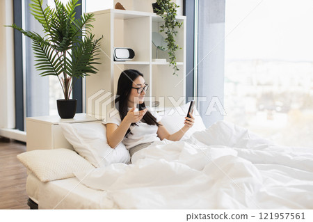 Asian woman in her 20s using smartphone for online video call while sitting on bed. Relaxed atmosphere with indoor plants and modern decor. 121957561