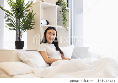 Asian woman in modern bedroom working on laptop, surrounded by plants and natural light, creating calm and productive atmosphere. Asian woman in modern bedroom working on laptop, surrounded by plants and natural light, creating calm and productive atmosphere. 121957583