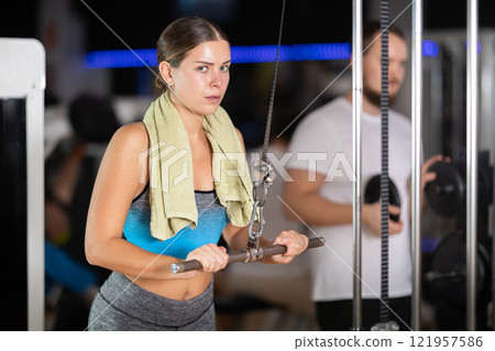 Girl in gym performs exercises for arm muscles using cable crossover machine and pull weighted rope 121957586