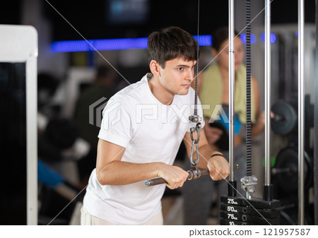 Guy in gym performs exercises for arm muscles using cable crossover machine and pull weighted rope Guy in gym performs exercises for arm muscles using cable crossover machine and pull weighted rope 121957587