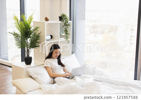Asian woman in her 20s using laptop in cozy bedroom setting. Relaxed and focused atmosphere with natural daylight and indoor plants adding calmness to home workspace. 121957588