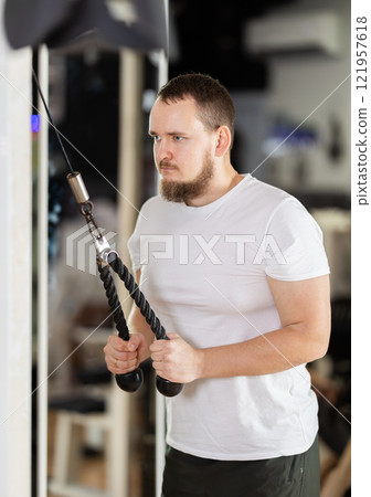 Man in gym performs exercises for arm muscles using cable crossover machine and pull weighted rope Man in gym performs exercises for arm muscles using cable crossover machine and pull weighted rope 121957618