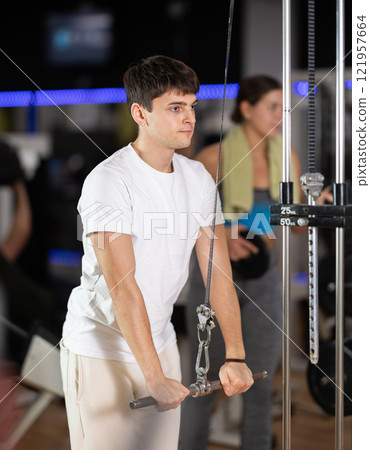 Guy in gym performs exercises for arm muscles using cable crossover machine and pull weighted rope Guy in gym performs exercises for arm muscles using cable crossover machine and pull weighted rope 121957664