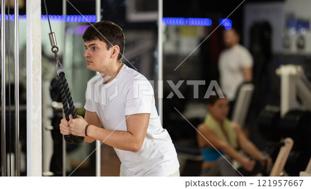 Guy in gym performs exercises for arm muscles using cable crossover machine and pull weighted rope Guy in gym performs exercises for arm muscles using cable crossover machine and pull weighted rope 121957667