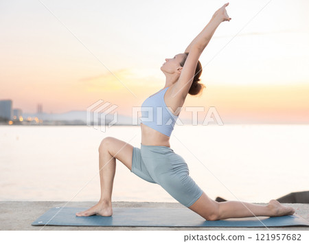 Sporty energetic female teacher doing exercises on pilates mat and demonstrating variation of warrior pose of yoga on beach at sunset. Fitness concept 121957682
