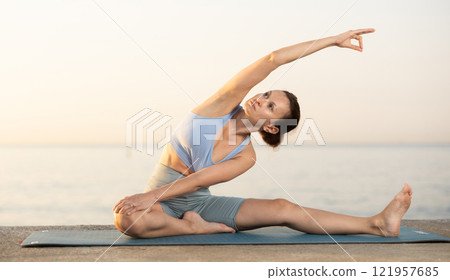 Near sea during sunset, girl performs variations of Janu Shirshasana 121957685