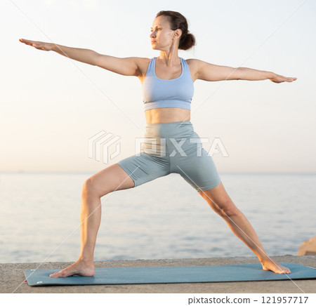 Near sea during sunset, girl performs variations of virabhadrasana 121957717