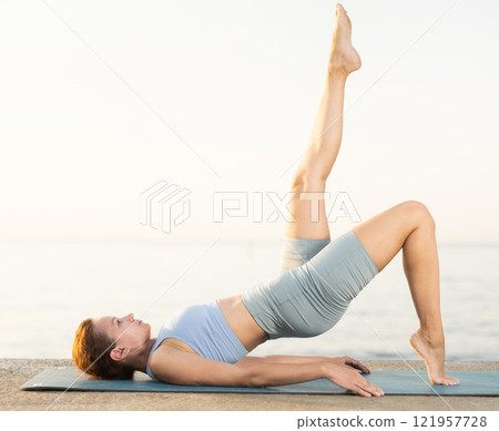 Near sea during sunset, girl performs Setu Badha Sarvangasana 121957728