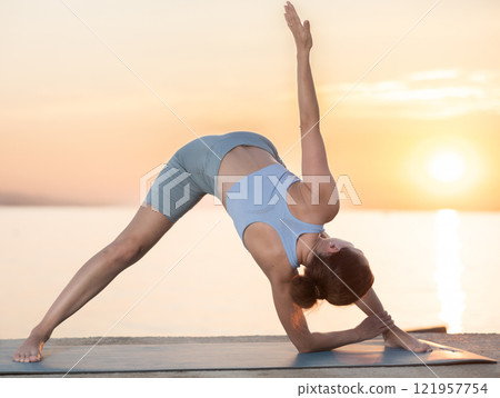 Near sea during sunset, girl performs variations of Himavanasana 121957754