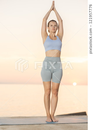 Near sea during sunset, girl performs Tiryaka Tadasana 121957776
