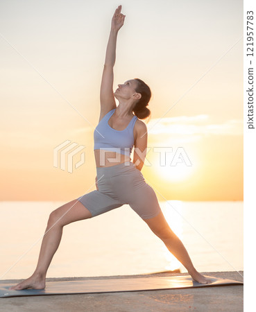 Sporty woman practicing yoga outside, doing relaxing stretching workout for body flexibility on seashore at sunrise Sporty woman practicing yoga outside, doing relaxing stretching workout for body flexibility on seashore at sunrise 121957783