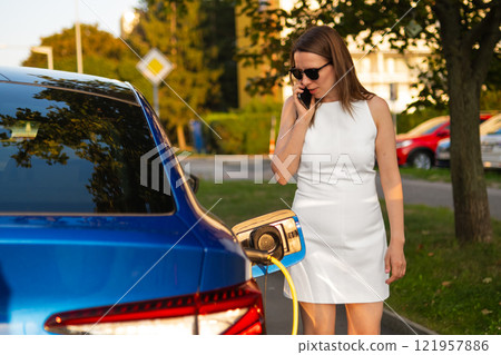 Woman in a white dress stands beside a charging electric car, talking on her mobile phone while parked outdoors. Woman in a white dress stands beside a charging electric car, talking on her mobile phone while parked outdoors. 121957886