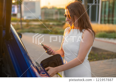 Woman in a white dress uses her smartphone while charging her electric vehicle in the street Woman in a white dress uses her smartphone while charging her electric vehicle in the street 121957887