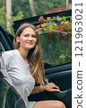 A cheerful young woman with a bright smile poses by a parked car, surrounded by vibrant greenery and nature 121963021