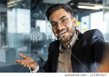 Video call, businessman smiling looking into smartphone camera. Man at workplace inside office in business suit working, using smartphone app for remote conversation. 121963427