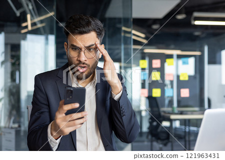 Man upset and disappointed holding phone in hands inside office. Businessman received online message with bad news on smartphone 121963431