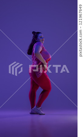 Side view photo of young plus size woman dressed red sportswear posing confidently against purple studio background in vibrant neon light. Side view photo of young plus size woman dressed red sportswear posing confidently against purple studio background in vibrant neon light. 121967649