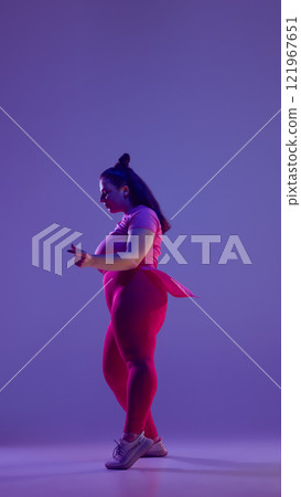 Plus size young active woman dressed red sportswear dancing hip-hop in motion against purple studio background in vibrant neon light. Plus size young active woman dressed red sportswear dancing hip-hop in motion against purple studio background in vibrant neon light. 121967651