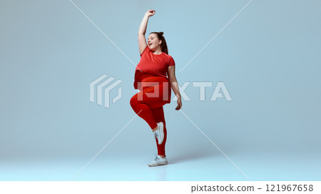 Smiling chubby woman wearing red t-shirt, red leggings, white sneakers, standing mid-dance with hands high against beige studio background. Smiling chubby woman wearing red t-shirt, red leggings, white sneakers, standing mid-dance with hands high against beige studio background. 121967658