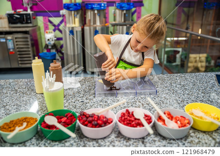 Little girl learning how to cook in a cooking class. Handmade dessert. Making chocolate candy, Child filling heart shaped mold with chocolate Little girl learning how to cook in a cooking class. Handmade dessert. Making chocolate candy, Child filling heart shaped mold with chocolate 121968479