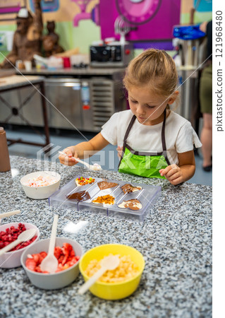Little girl learning how to cook in a cooking class. Handmade dessert. Making chocolate candy, Child filling heart shaped mold with chocolate 121968480