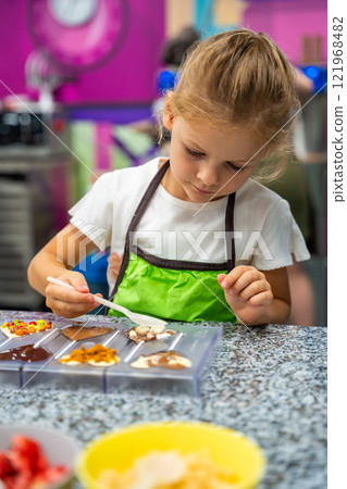 Little girl learning how to cook in a cooking class. Handmade dessert. Making chocolate candy, Child filling heart shaped mold with chocolate Little girl learning how to cook in a cooking class. Handmade dessert. Making chocolate candy, Child filling heart shaped mold with chocolate 121968482